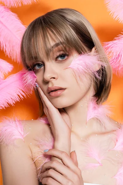 Jeune femme avec des perles décoratives en maquillage toucher la joue près de plumes roses isolées sur orange — Photo de stock