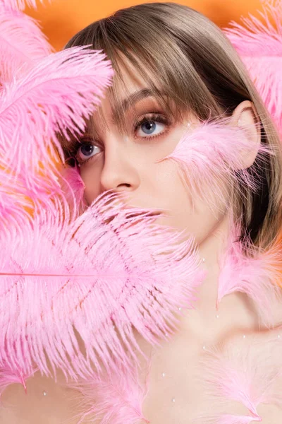 Mujer joven con cuentas decorativas en maquillaje mirando hacia otro lado a través de plumas rosadas aisladas en naranja - foto de stock