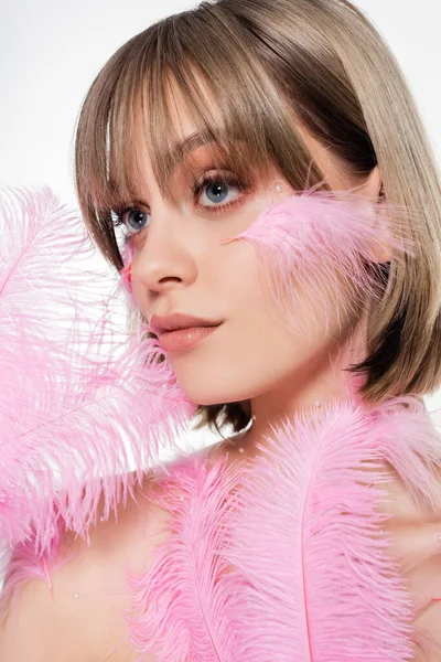 Bonita mujer con cuentas decorativas en el cuerpo mirando hacia otro lado y sosteniendo plumas rosadas aisladas en blanco - foto de stock