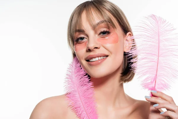 Positive young woman with hydrogel eye patches holding pink feathers isolated on white — Foto stock