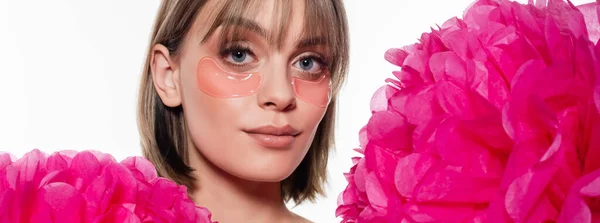 Mujer joven con parches de ojo de hidrogel cerca de flores de color rosa brillante aislado en blanco, pancarta - foto de stock