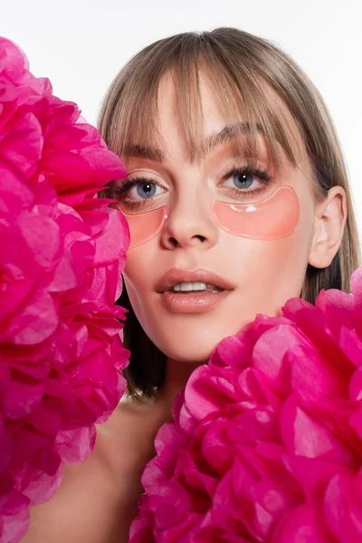 Giovane donna con macchie oculari idrogel e occhi azzurri vicino a fiori rosa brillante isolati su bianco — Foto stock