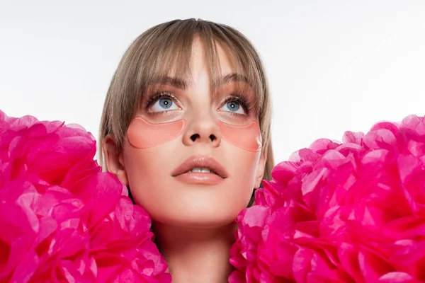 Giovane donna con macchie oculari idrogel guardando verso l'alto vicino a fiori rosa brillante isolati su bianco — Foto stock