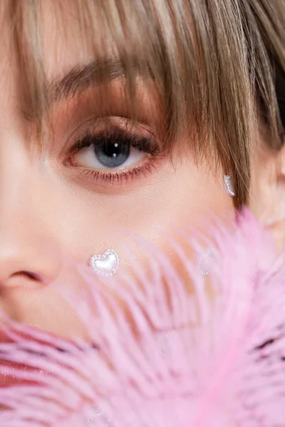 Vista recortada de la mujer joven con elementos de forma de corazón nacreo en la mejilla cerca de la pluma rosa - foto de stock