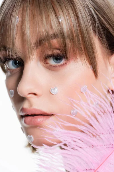 Close-up de mulher jovem com nacreous elementos de forma do coração em bochechas perto de penas rosa isolado em branco — Fotografia de Stock