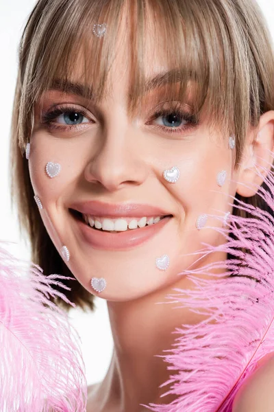 Primer plano de mujer feliz con elementos de forma de corazón nacreo en las mejillas cerca de plumas rosadas aisladas en blanco - foto de stock