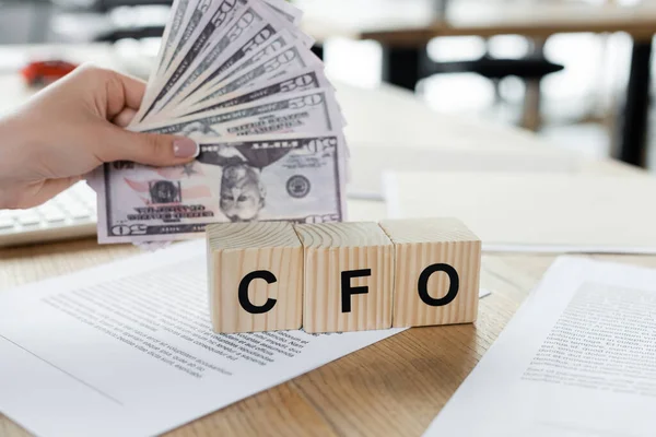 Cropped view of woman holding dollars near cubes with cfo lettering on papers on desk — Stock Photo