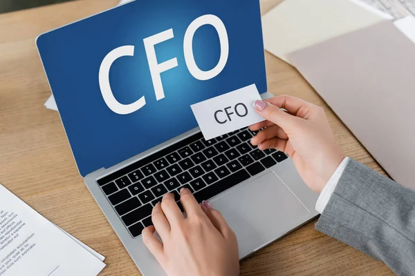 Cropped view of woman holding paper and using laptop with cfo lettering on screen — Stock Photo