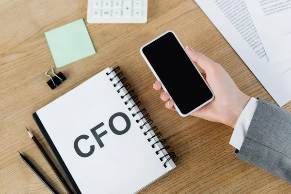 Top view of woman holding smartphone with blank screen near notebook with cfo lettering and stationery — Stock Photo