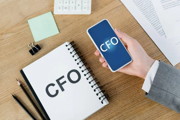 Top view of woman holding smartphone with cfo lettering on screen near notebook and stationery — Stock Photo