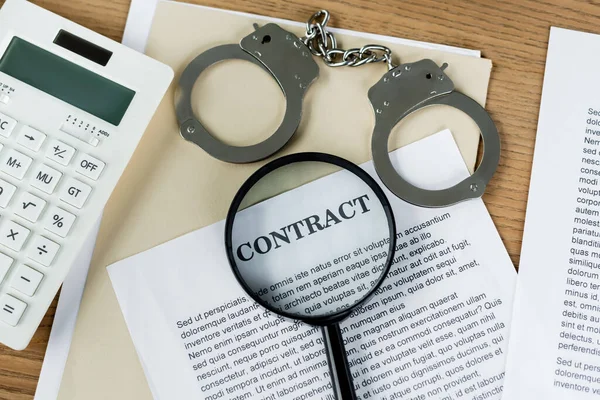 Top view of magnifier on papers with contract lettering near calculator and handcuffs — Stock Photo