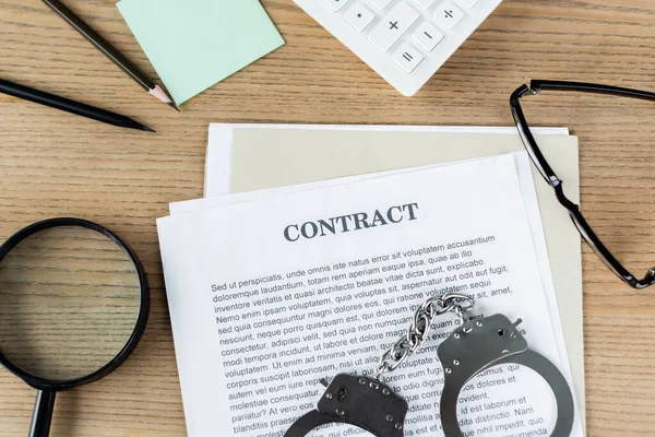 Top view of handcuffs on contract near magnifier, glasses and stationery — Stock Photo