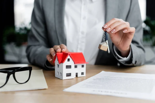 Ausgeschnittene Ansicht einer Frau, die Schlüsselanhänger mit Schlüssel in der Nähe des Hausmodells und Dokumente auf dem Schreibtisch hält — Stockfoto