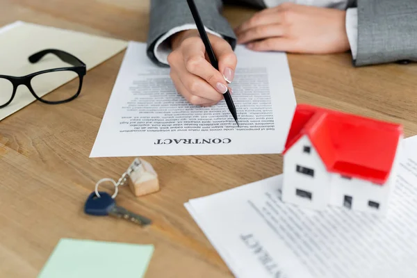 Vue recadrée de femme signant contrat près de porte-clés avec clé et modèle de maison sur le bureau — Photo de stock