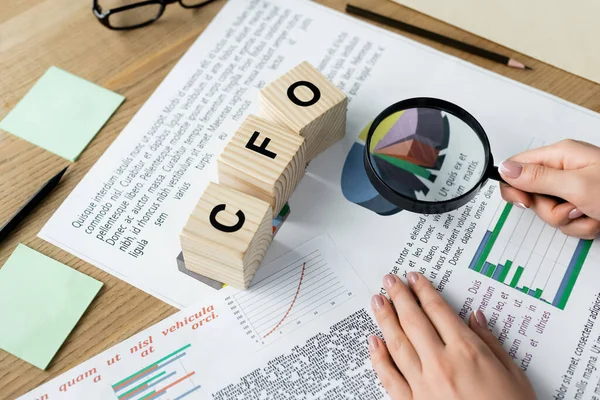 Cropped view of woman holding magnifier above charts near cubes with cfo lettering on desk — Stock Photo