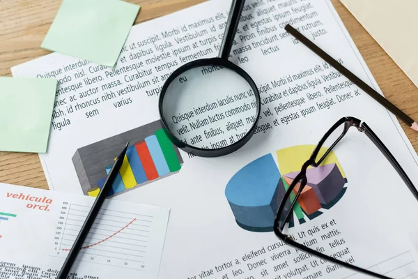 Top view of magnifier and glasses on charts near stationery on desk — Stock Photo