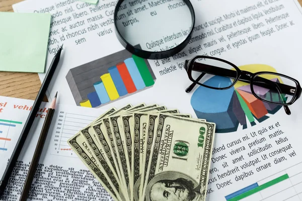 Top view of magnifier and glasses on charts near dollar banknotes on desk — Stock Photo