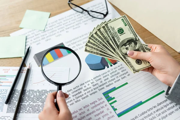 Top view of tax inspector holding magnifier and glasses above charts and dollar banknotes — Stock Photo