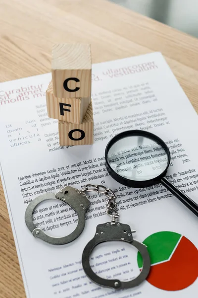 Magnifying glass near cubes with cfo letters, charts and handcuffs on desk — Stock Photo