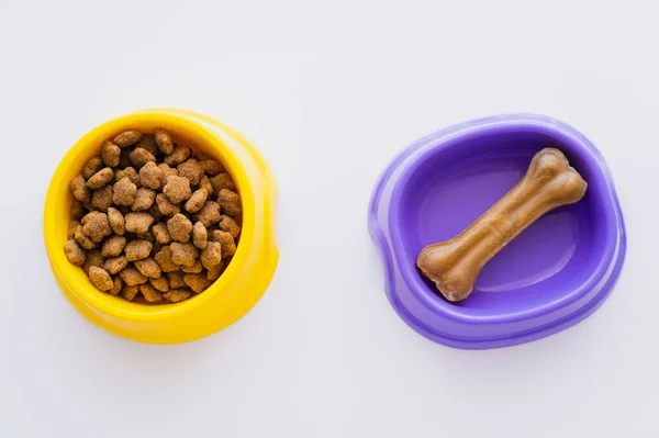 Vista superior de las golosinas de mascotas en forma de hueso en un tazón cerca de alimentos secos para mascotas aislados en blanco - foto de stock