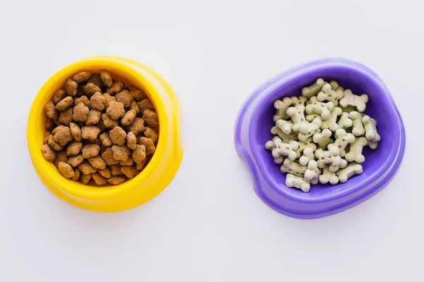 Vista superior de las golosinas de mascotas en forma de hueso cerca de alimentos secos para mascotas en cuencos aislados en blanco - foto de stock