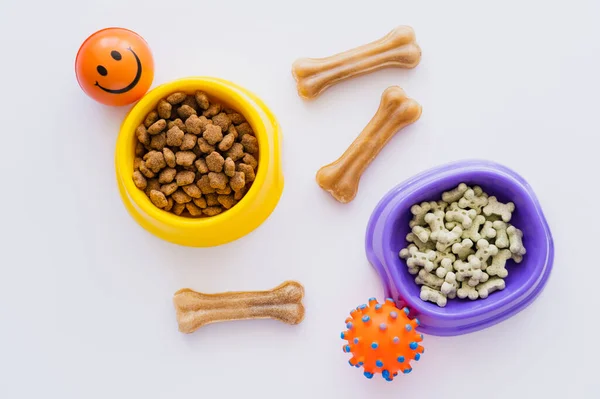 Vista superior de biscoitos de estimação em forma de osso perto de alimentos secos para animais de estimação em tigelas e brinquedos de borracha em branco — Fotografia de Stock