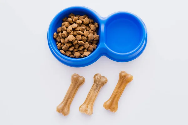 Vista superior de las golosinas para mascotas en forma de hueso cerca de un tazón con agua y comida seca para mascotas aislada en blanco - foto de stock