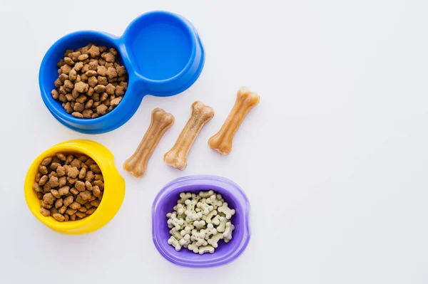Vista superior de las golosinas de mascotas en forma de hueso cerca de cuencos con agua y alimentos secos para mascotas aislados en blanco - foto de stock