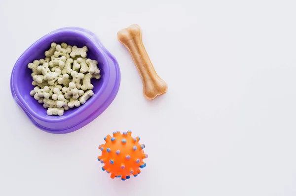 Vue du dessus des biscuits pour animaux de compagnie en forme d'os dans un bol violet près d'une balle en caoutchouc isolée sur blanc — Photo de stock