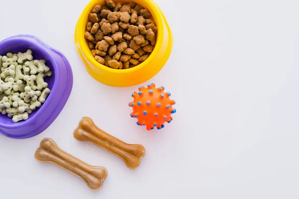 Vue de dessus des biscuits pour animaux en forme d'os et de la nourriture sèche dans des bols près de boule de caoutchouc isolé sur blanc — Photo de stock