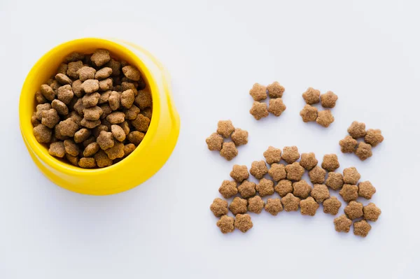 Vista superior de forma de pata hecha de comida seca para mascotas cerca de un tazón aislado en blanco - foto de stock