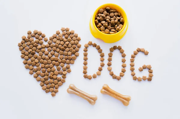 Vue de dessus de nourriture sèche pour animaux de compagnie en forme de coeur près de lettrage de chien et les os isolés sur blanc — Photo de stock