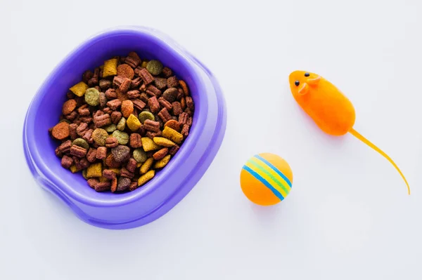 Вид сверху фиолетовой пластиковой миски с сухим кормом для домашних животных рядом с резиновой игрушечной мышью и шаром, изолированным на белом — стоковое фото