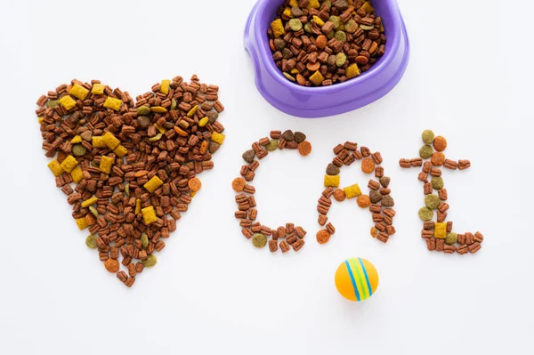 Vue du dessus du mot coeur et chat fait de nourriture sèche pour animaux de compagnie près du bol et de la balle isolé sur blanc — Photo de stock