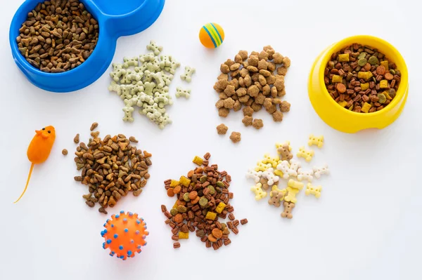 Vista superior de diferentes alimentos secos y deliciosas golosinas para mascotas cerca de juguetes y tazones aislados en blanco - foto de stock