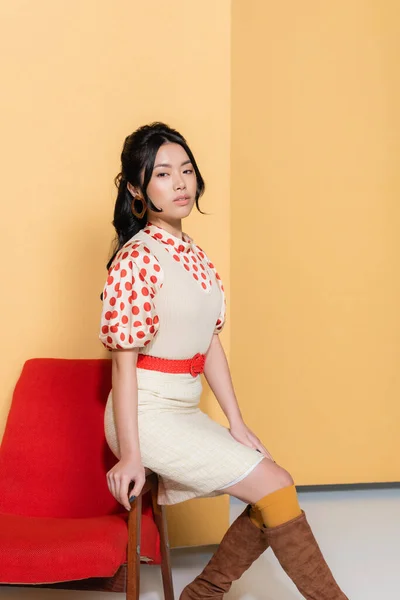 Modelo asiático de moda sentado en sillón retro sobre fondo naranja - foto de stock