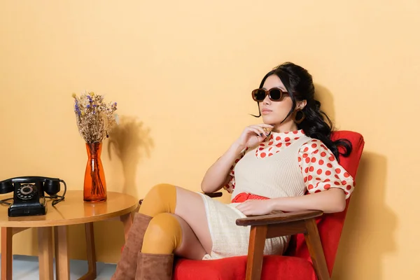 Mujer asiática de moda sentada en un sillón cerca del teléfono y flores en la mesa de café sobre fondo naranja - foto de stock