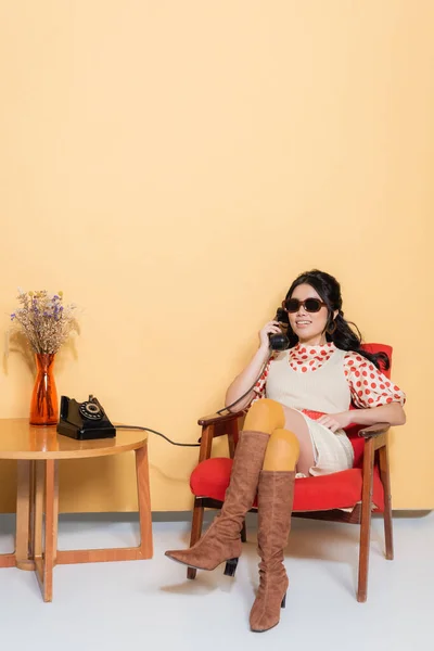 Souriant asiatique femme en lunettes de soleil et des vêtements vintage parler au téléphone tout en étant assis sur un fauteuil sur fond orange — Photo de stock