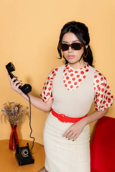 Elegante mujer asiática en gafas de sol sosteniendo el teléfono sobre fondo naranja - foto de stock