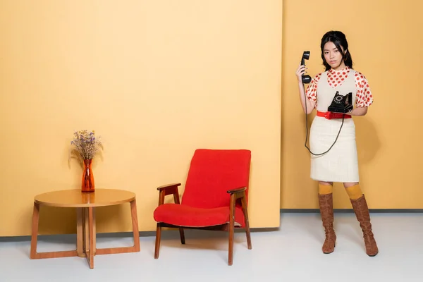Pleine longueur de modèle asiatique élégant tenant le téléphone près du fauteuil et de la table basse sur fond orange — Photo de stock