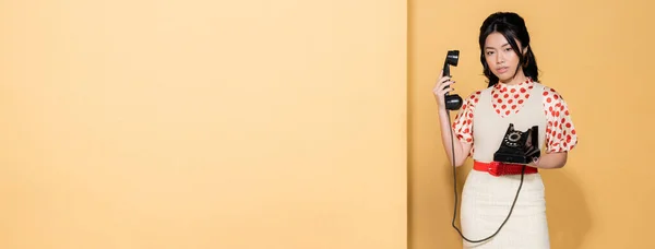 Joven modelo asiático en ropa retro sosteniendo teléfono sobre fondo naranja, bandera - foto de stock