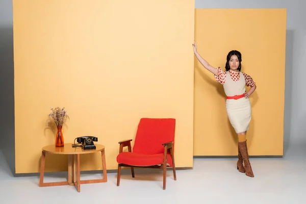 Moda asiática modelo posando perto de poltrona e mesa de café no fundo laranja — Fotografia de Stock