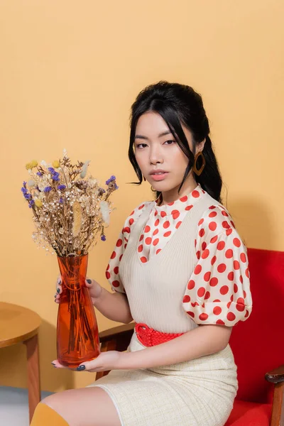 Stilvolle asiatische Frau hält Vase mit Blumen, während sie auf einem Sessel auf orangefarbenem Hintergrund sitzt — Stockfoto