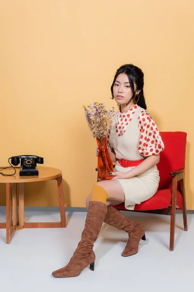 Jeune modèle asiatique en vêtements vintage tenant des fleurs tout en étant assis sur un fauteuil sur fond orange — Photo de stock