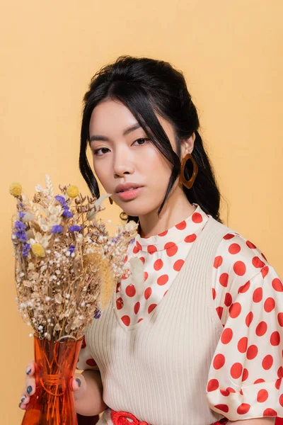 Портрет молодой азиатской модели в винтажной одежде, держащей вазу с цветами, изолированными на оранжевом — стоковое фото