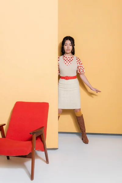 Elegante mujer asiática en ropa retro mirando a la cámara cerca de sillón sobre fondo naranja - foto de stock