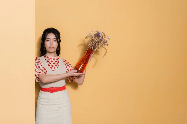 Elegante modelo asiático sosteniendo jarrón con flores sobre fondo naranja - foto de stock