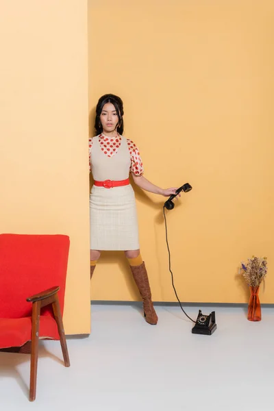 Полная длина стильной азиатской модели с телефоном рядом с креслом и цветами на оранжевом фоне — стоковое фото