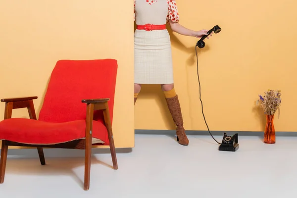 Vista recortada del modelo en ropa vintage sosteniendo el teléfono cerca de flores y sillón sobre fondo naranja - foto de stock