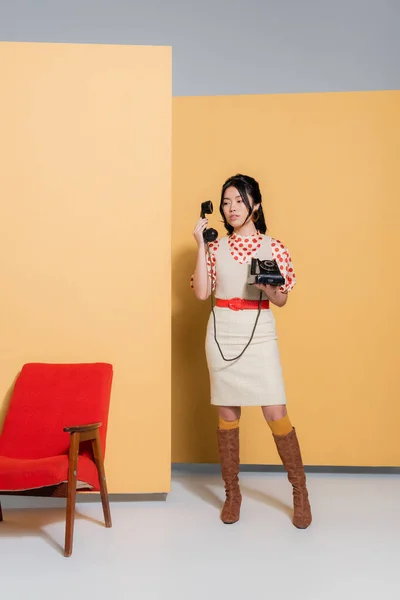 Trendy asiatische Frau in Vintage-Kleidung hält Telefon in der Nähe Sessel auf orangefarbenem Hintergrund — Stockfoto